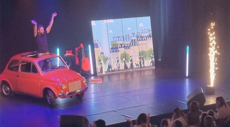 Une petite voiture rouge sur scène avec du public devant qui applaudit dans le cadre d'un spectacle pour enfant